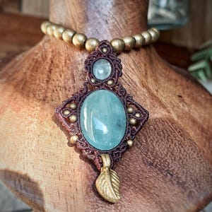 May include: A handcrafted necklace featuring a large, oval, light blue gemstone pendant. The pendant is intricately woven with brown cord and adorned with small gold beads. A smaller, round gemstone sits above the larger one, and a gold leaf charm hangs below.
