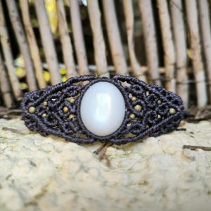 May include: A handmade macrame bracelet featuring a central oval white stone. The dark brown macrame is accented with small, gold-colored beads. The bracelet is displayed on a textured, light-colored surface.
