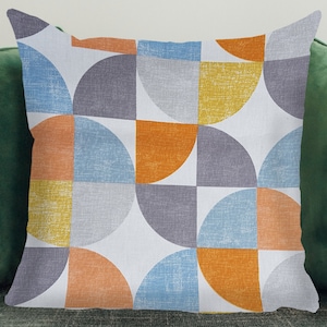 May include: Square cushion with a geometric pattern. The design features semi-circles in shades of orange, yellow, blue, and grey against a white background. The cushion is placed on a green surface.