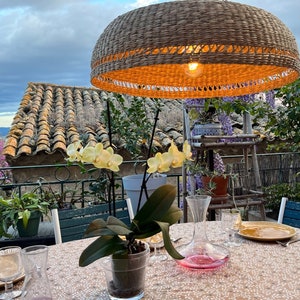 May include: A table setting with a wicker pendant light hanging above, a white tablecloth, and a glass decanter with a pink liquid. There are two glasses, a plate, and a napkin on the table. A potted orchid plant is in the centre of the table.