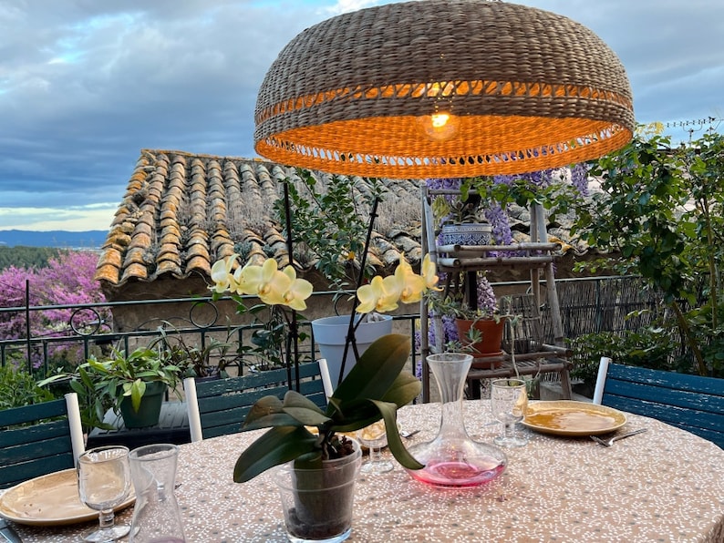 May include: A table setting with a wicker pendant light hanging above, a white tablecloth, and a glass decanter with a pink liquid. There are two glasses, a plate, and a napkin on the table. A potted orchid plant is in the centre of the table.