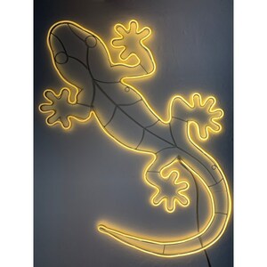 May include: A yellow neon light shaped like a gecko lizard. The light is mounted on a wall and is illuminated.