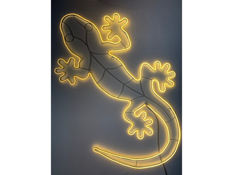 May include: A yellow neon light shaped like a gecko lizard. The light is mounted on a wall and is illuminated.