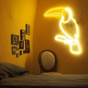 May include: A yellow neon toucan light fixture mounted on a yellow wall. The toucan is perched on a branch. There is a bed with a gray duvet and a chair in the background.