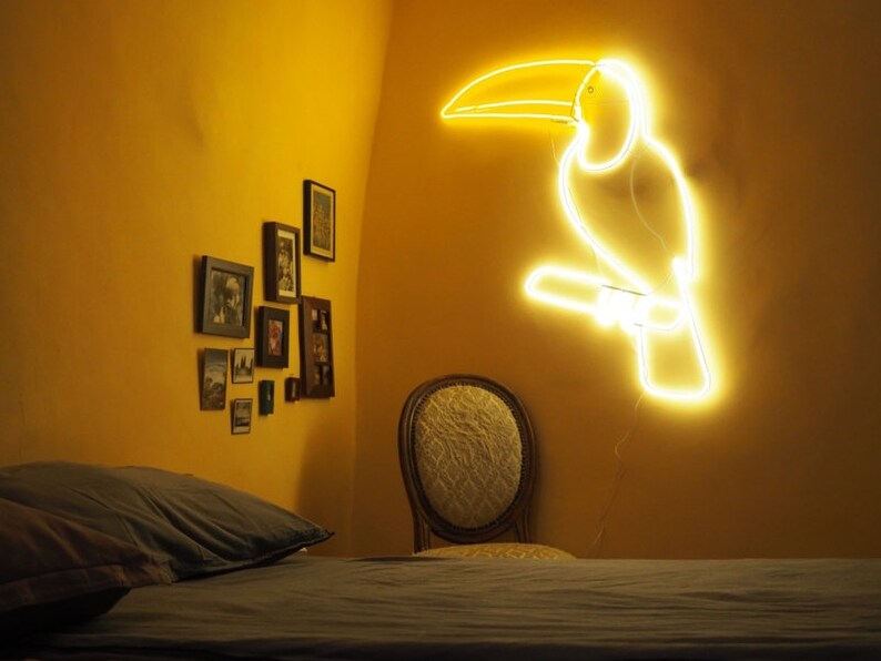 May include: A yellow neon toucan light fixture mounted on a yellow wall. The toucan is perched on a branch. There is a bed with a gray duvet and a chair in the background.