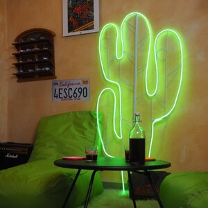 May include: A green neon cactus light sculpture on a wall with a green beanbag chair, a small table, and a glass of liquid. The wall also has a license plate that reads "JUL California 4ESC690".