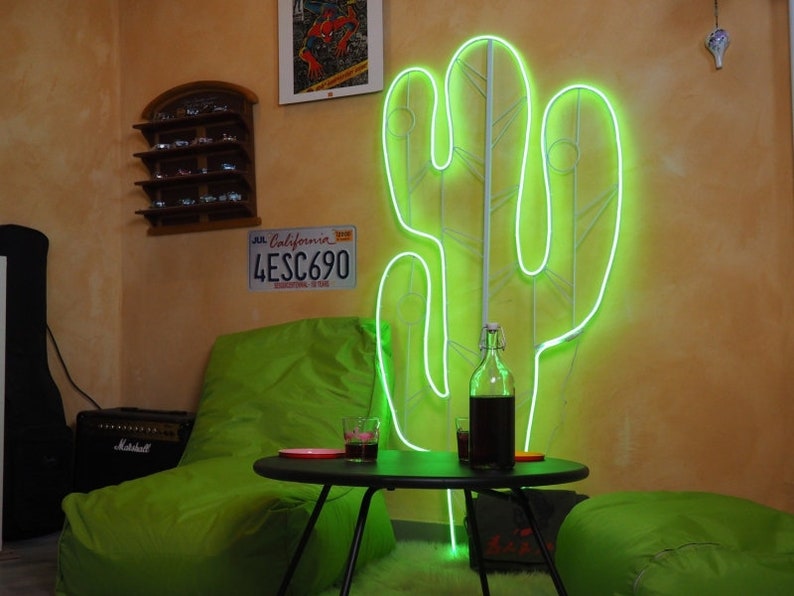 May include: A green neon cactus light sculpture on a wall with a green beanbag chair, a small table, and a glass of liquid. The wall also has a license plate that reads "JUL California 4ESC690".