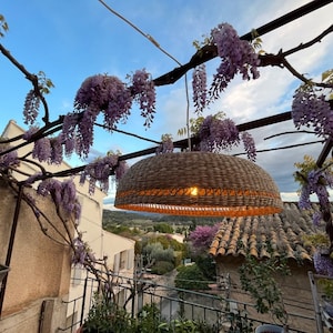 May include: A woven wicker pendant light hangs from a trellis covered in purple wisteria flowers. The light is illuminated and casts a warm glow. The background shows a view of a tiled roof and a green hillside.