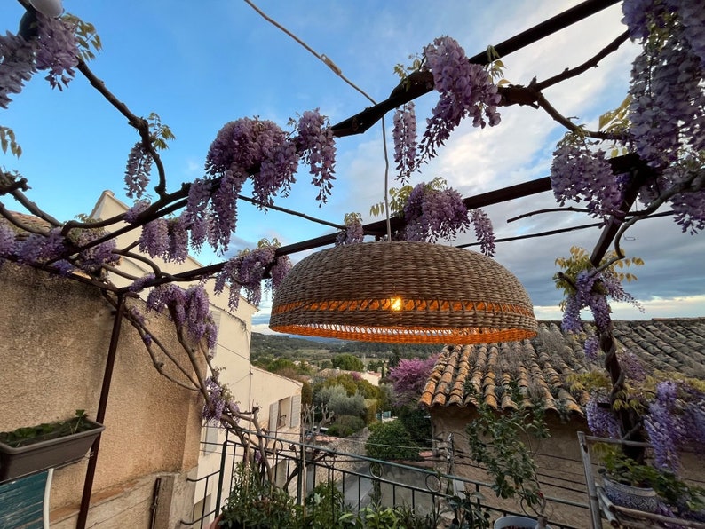 May include: A woven wicker pendant light hangs from a trellis covered in purple wisteria flowers. The light is illuminated and casts a warm glow. The background shows a view of a tiled roof and a green hillside.