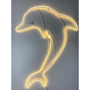 May include: A white neon dolphin sign with a yellow glow. The dolphin is jumping out of the water and has a curved tail.