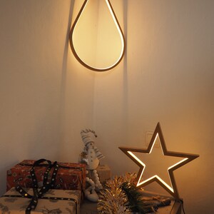 May include: A wooden teardrop-shaped light fixture hangs from the ceiling, illuminating a table with wrapped gifts, a snowman figurine, and a wooden star-shaped light fixture. The table is decorated with gold tinsel.