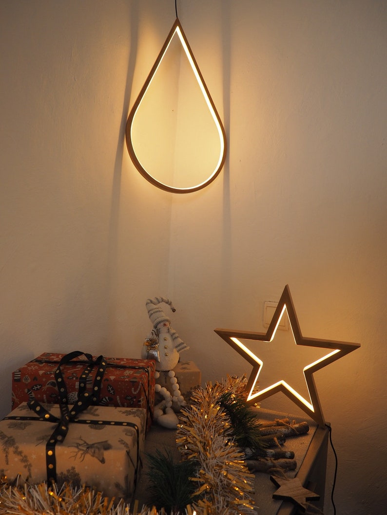 May include: A wooden teardrop-shaped light fixture hangs from the ceiling, illuminating a table with wrapped gifts, a snowman figurine, and a wooden star-shaped light fixture. The table is decorated with gold tinsel.
