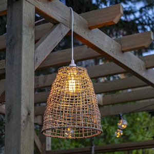 Puede incluir: Una lámpara colgante de mimbre tejido cuelga de una pérgola de madera. La luz está encendida e ilumina el área circundante con un brillo cálido.