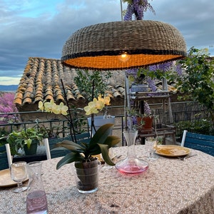 May include: A wicker pendant light hangs over a table set for dinner on a patio. The table has a white tablecloth with a floral pattern and is set with two glasses, a decanter, and a plate. There are plants and flowers in pots on the table and in the background.