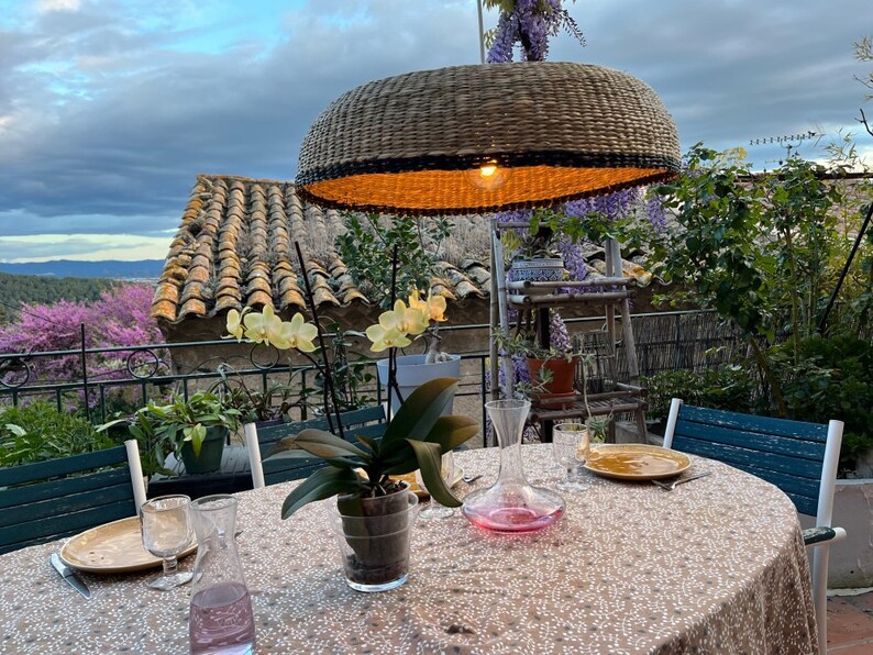 May include: A wicker pendant light hangs over a table set for dinner on a patio. The table has a white tablecloth with a floral pattern and is set with two glasses, a decanter, and a plate. There are plants and flowers in pots on the table and in the background.