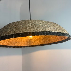 May include: A woven pendant light with a black trim and a warm white light bulb. The light fixture is made of natural materials and has a rustic, bohemian style.