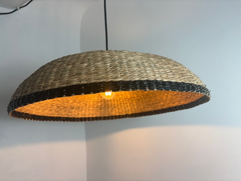 May include: A woven pendant light with a black trim and a warm white light bulb. The light fixture is made of natural materials and has a rustic, bohemian style.