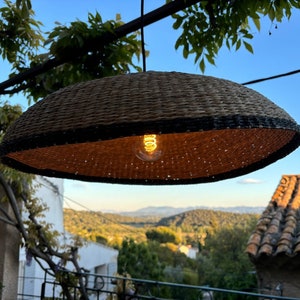 May include: A woven wicker pendant light with a black trim hangs from a branch, illuminating the surrounding area with a warm glow. The light is suspended over a view of a hillside with trees and a tiled roof in the distance.