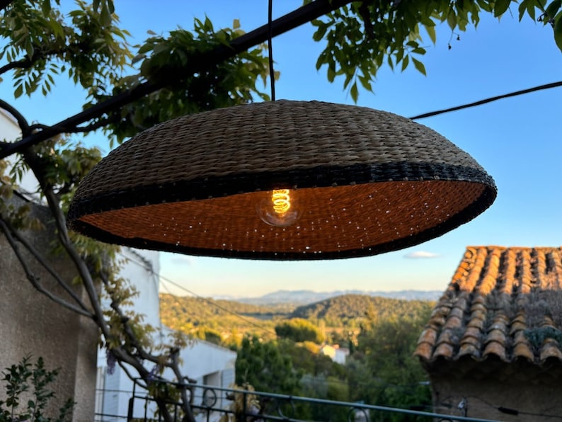 May include: A woven wicker pendant light with a black trim hangs from a branch, illuminating the surrounding area with a warm glow. The light is suspended over a view of a hillside with trees and a tiled roof in the distance.