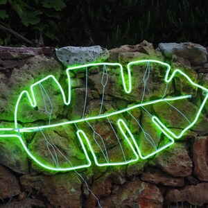 May include: A green neon light shaped like a tropical leaf is mounted on a stone wall. The light is on and glowing brightly.