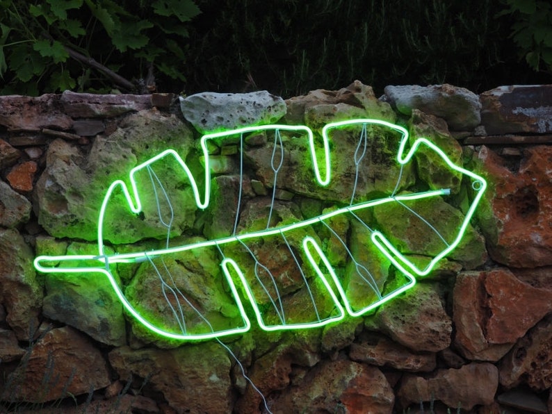 May include: A green neon light shaped like a tropical leaf is mounted on a stone wall. The light is on and glowing brightly.