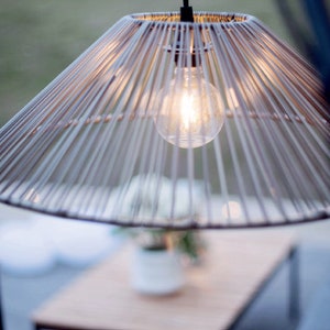 Può includere: Un paralume intrecciato di colore marrone chiaro con una lampadina rotonda e trasparente appesa a un filo nero. Il paralume è appeso sopra un tavolo di legno.