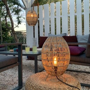 May include: A wicker pendant light hangs above a wicker floor lamp with a light bulb illuminating the space. The lamp is on a woven rug in front of a white slatted fence and a dark gray couch with pillows.