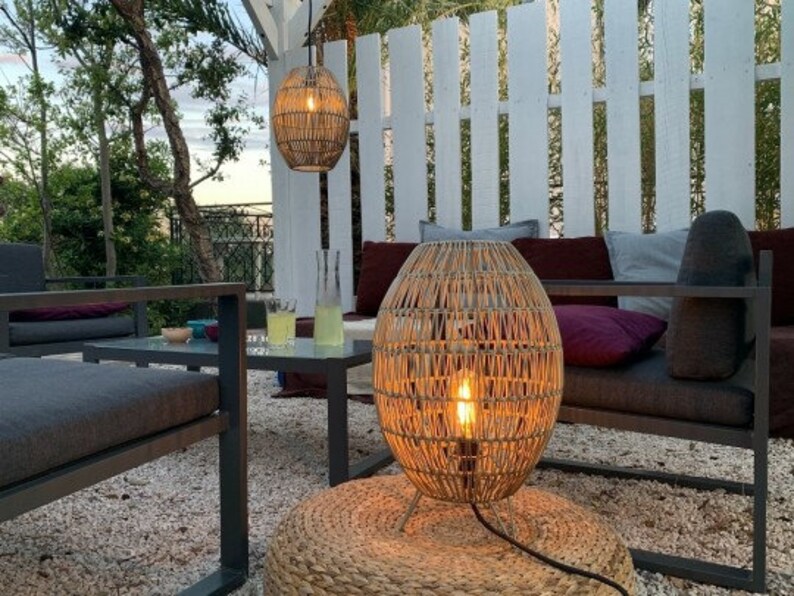 May include: A wicker pendant light hangs above a wicker floor lamp with a light bulb illuminating the space. The lamp is on a woven rug in front of a white slatted fence and a dark gray couch with pillows.