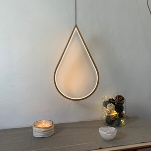 May include: A white teardrop-shaped pendant light fixture with a wooden frame hangs from the ceiling. The light is on and illuminates the room. There are two candles and a glass vase with decorative balls on a table below the light.