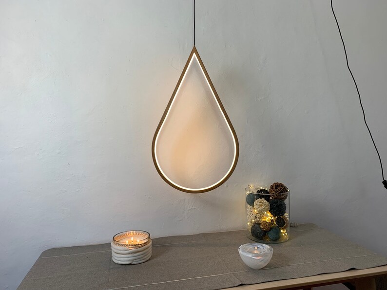 May include: A white teardrop-shaped pendant light fixture with a wooden frame hangs from the ceiling. The light is on and illuminates the room. There are two candles and a glass vase with decorative balls on a table below the light.