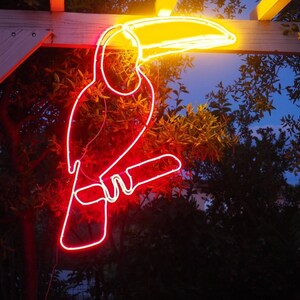 May include: A neon toucan sign with a red body and yellow beak, hanging from a wooden pergola. The sign is lit up and glowing brightly against a dark background.