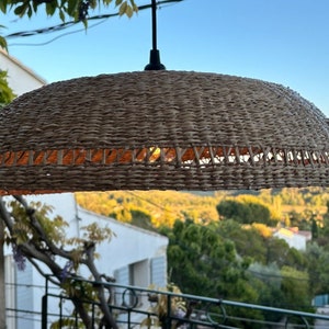 May include: A woven wicker pendant light fixture in a natural brown colour. The light fixture is hanging from a black cord and is illuminated with a warm white light.