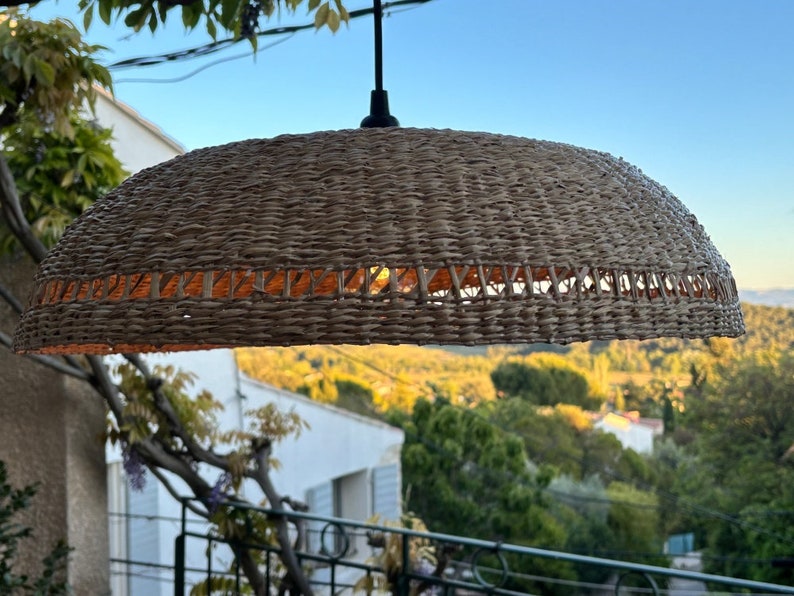 May include: A woven wicker pendant light fixture in a natural brown colour. The light fixture is hanging from a black cord and is illuminated with a warm white light.