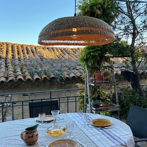 May include: A wicker pendant light hangs over a table set for dinner on a patio. The table has a white tablecloth with a floral pattern and a jug with a red and white design. The patio has a view of a tiled roof and a lush green garden.