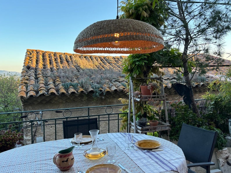 May include: A wicker pendant light hangs over a table set for dinner on a patio. The table has a white tablecloth with a floral pattern and a jug with a red and white design. The patio has a view of a tiled roof and a lush green garden.