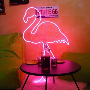 May include: Pink neon flamingo light sculpture with a black table and two glasses of dark liquid in front of it. The neon light is on and illuminates the room. The background is a peach colored wall with a sign that reads "Arizona Route 66".