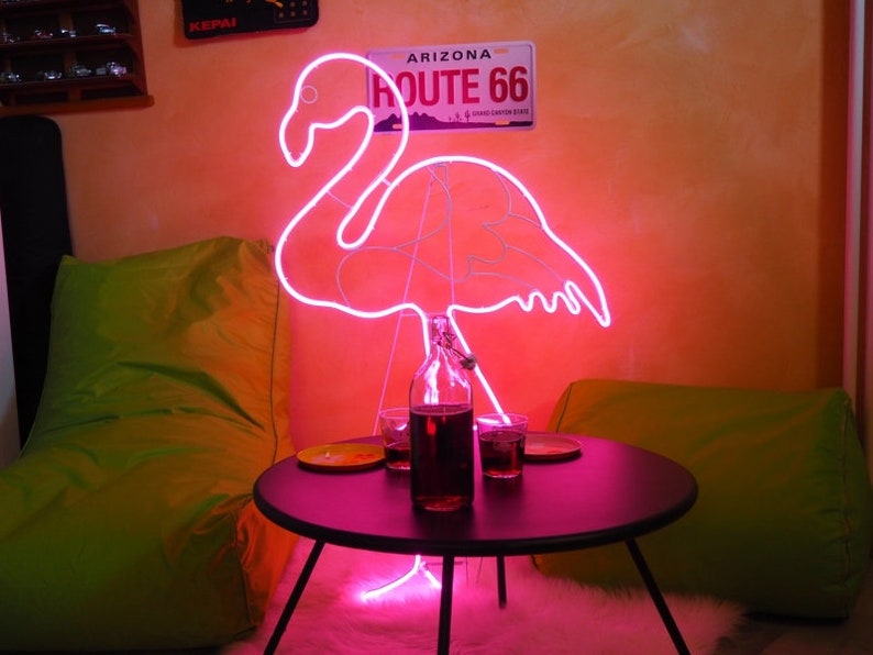 May include: Pink neon flamingo light sculpture with a black table and two glasses of dark liquid in front of it. The neon light is on and illuminates the room. The background is a peach colored wall with a sign that reads "Arizona Route 66".