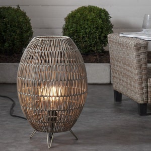 May include: A wicker outdoor floor lamp with a round, woven shade and a light bulb inside. The lamp is on a metal stand with three legs and is plugged into an electrical outlet.