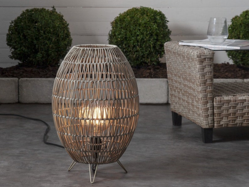 May include: A wicker outdoor floor lamp with a round, woven shade and a light bulb inside. The lamp is on a metal stand with three legs and is plugged into an electrical outlet.