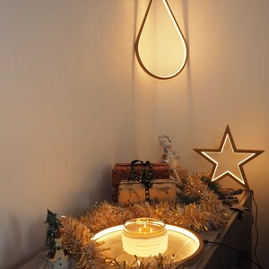 May include: A wooden teardrop-shaped light fixture hangs from the ceiling, illuminating a table with a lit candle, a snowman figurine, and gold tinsel. A star-shaped light fixture is on the wall behind the table.