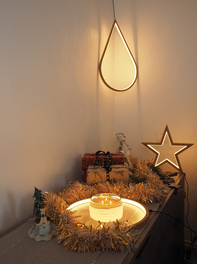 May include: A wooden teardrop-shaped light fixture hangs from the ceiling, illuminating a table with a lit candle, a snowman figurine, and gold tinsel. A star-shaped light fixture is on the wall behind the table.