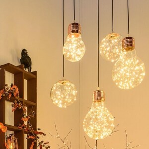 Puede incluir: Cinco lámparas colgantes de tono cobre con bombillas de vidrio transparente llenas de luces blancas parpadeantes. Las bombillas cuelgan de un techo blanco contra una pared blanca.