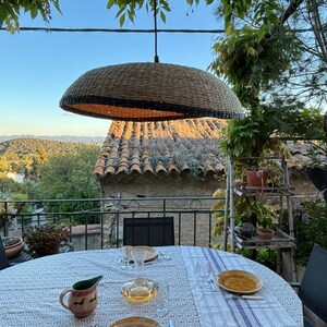May include: A table set for a meal on a balcony with a view of a mountain range in the distance. The table has a white tablecloth with a blue and white pattern. There is a wicker lampshade hanging above the table.