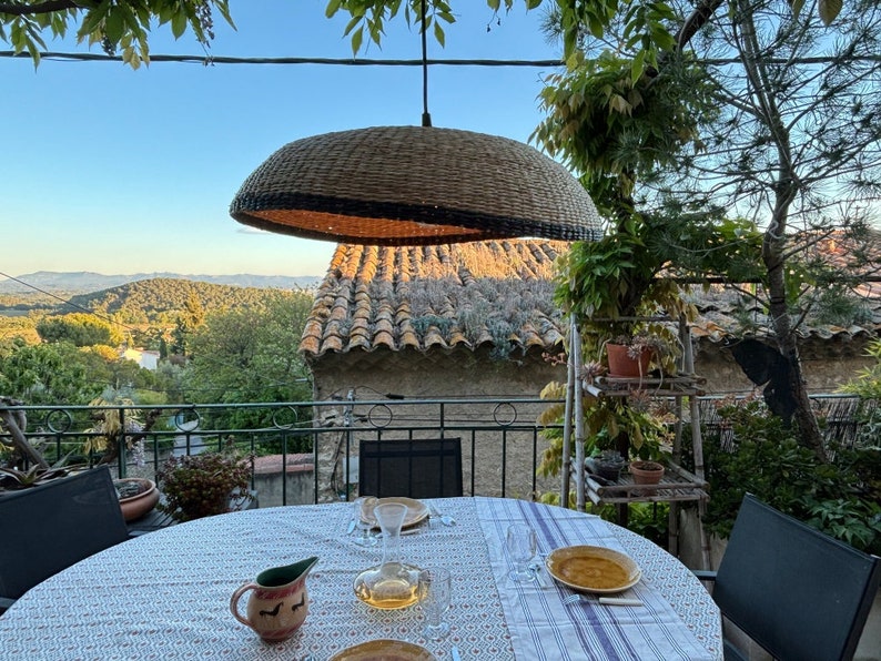 May include: A table set for a meal on a balcony with a view of a mountain range in the distance. The table has a white tablecloth with a blue and white pattern. There is a wicker lampshade hanging above the table.
