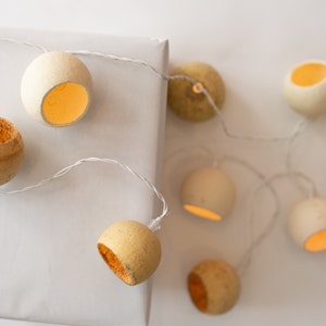 Può includere: Una ghirlanda di luci con un filo bianco e lampadine di carta marrone chiaro. Le lampadine sono accese ed emettono una luce calda gialla. Le luci sono infilate attorno a una scatola regalo bianca.