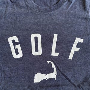 May include: Navy blue t-shirt with the word "GOLF" in large white letters. Below the word is a white graphic of a peninsula. The shirt has a crew neck and is made of a soft material.