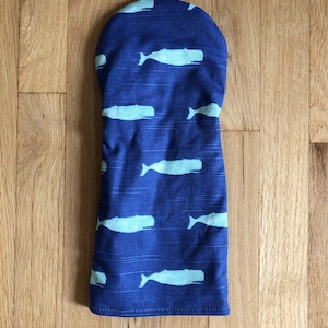 May include: A blue golf club head cover with a pattern of light blue whales. The cover is made of fabric and has a rounded top. The whales are swimming horizontally across the cover.