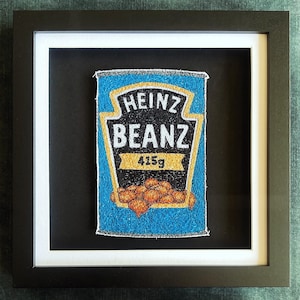 May include: Framed artwork of a Heinz Beanz can. The can is blue with the Heinz logo and the word "BEANZ". The artwork includes the text "415g" and a depiction of baked beans. The artwork is framed in a black frame.