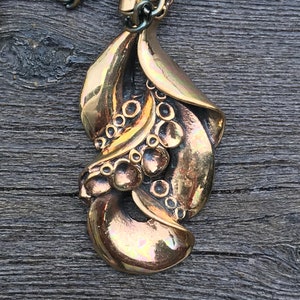 May include: A bronze pendant necklace with a detailed design of stylized leaves and berries. The pendant is attached to a silver chain.