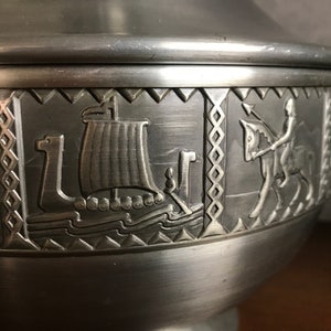 Puede incluir: Un taz&oacute;n de servir de peltre plateado con tapa, que presenta un dise&ntilde;o en relieve de un barco vikingo, un jinete y un patr&oacute;n geom&eacute;trico.